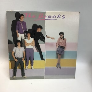 May include: Album cover for "The Breaks" featuring a band of six people. The album art has a retro aesthetic with pastel colors and the band's name in pink and purple. The band members are dressed in 1980s fashion.