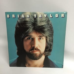 May include: A vintage vinyl record album cover featuring a portrait of Brian Taylor. The cover is blue with a close-up photo of Brian Taylor with long brown hair and a beard. The album title is printed in white letters at the top of the cover.