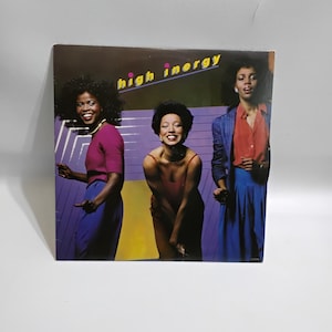 May include: Album cover featuring three women in vibrant 1970s attire. The album title "High Inergy" is in yellow text. The women are in shades of red, purple, and blue, with a purple and yellow background. The women are smiling.