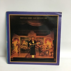 May include: A vintage vinyl record album cover featuring Emmylou Harris. The cover art depicts a woman in a black dress holding a guitar, standing on a stage in a saloon setting. The album title "Blue Kentucky Girl" is printed in white on a blue background.