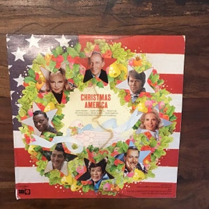 May include: A vintage vinyl record album cover featuring a festive wreath with stars and a variety of singers, including Bing Crosby, Glen Campbell, Peggy Lee, Nat King Cole, and Ella Fitzgerald. The album title is "Christmas America".