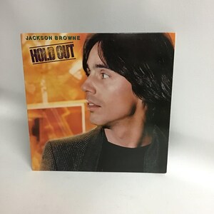 May include: A vinyl record album cover featuring a portrait of Jackson Browne. The album title "Hold Out" is printed in bold white letters on a black background.