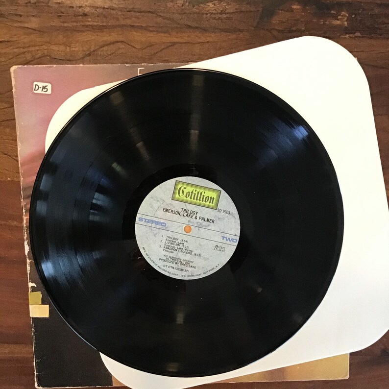 May include: A black vinyl record with a white label. The label has the text "Cotillion" and "Trilogy" in yellow. The text "Emerson, Lake & Palmer" is in black. The record is on a white turntable slipmat.
