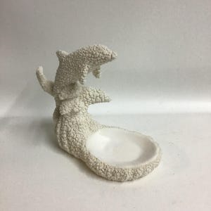 Könnte beinhalten: Weiße Keramik-Delfin-Schale mit strukturierter Oberfläche. Die Schale ist in Form von zwei Delfinen geformt, die aus dem Wasser springen. Die Schale hat eine kleine, runde Vertiefung in der Mitte.