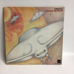 May include: A vintage vinyl record album cover featuring a drawing of a flying saucer with a red, orange, and yellow background. The album title is "Tailgunner" and the artist is "Jimmy McGriff".