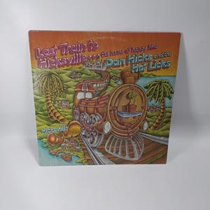 May include: Album cover for "Last Train to Hicksville" by Dan Hicks and the Hot Licks. The cover art features a cartoon train, palm trees, and the text "Last Train to Hicksville" and "the home of happy feet".
