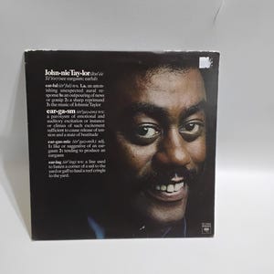 May include: Album cover featuring a smiling man with a mustache. The album title is "John-nie Tay-lor" with definitions of "earful", "ear-ga-sm", "ear-gas-mic", and "ear-ing" printed on the cover. The background is dark.
