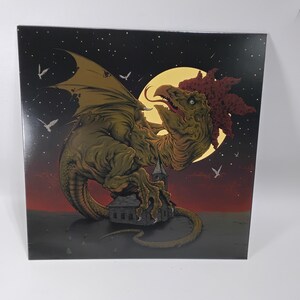 May include: A square print featuring a fantastical creature with dragon-like wings and a rooster-like head, set against a night sky with a full moon and a small church. The creature is primarily olive green with red accents, and the background transitions from black to red.