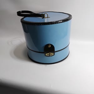 Könnte beinhalten: Eine hellblaue, zylindrische Handtasche mit glänzendem Finish. Die Tasche hat schwarze Verzierungen, einen schwarzen Griff und einen Verschluss. Die Handtasche hat einen Durchmesser von etwa 20 cm und ist 25 cm hoch.