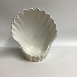 May include: White ceramic bowl shaped like a seashell, with a textured exterior. The bowl is designed to resemble a large seashell.