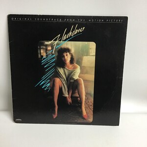 May include: A black vinyl record album cover with the title "Flashdance" in white text. The cover features a woman in a white sweater and red shoes sitting on a chair.