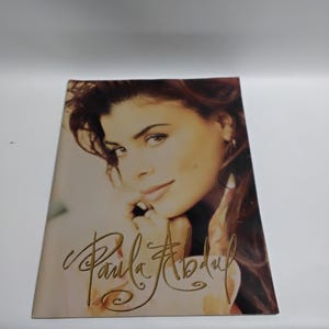 May include: A promotional item featuring a portrait of Paula Abdul. The image shows a woman with brown hair and a soft, sepia-toned background. The name "Paula Abdul" is written in a gold script font.