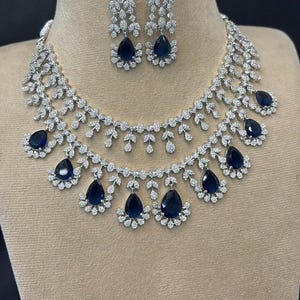 May include: A dazzling jewelry set featuring a necklace and matching earrings. The necklace and earrings are adorned with clear, sparkling stones and deep blue teardrop-shaped gems. The set is displayed on a neutral-toned mannequin.