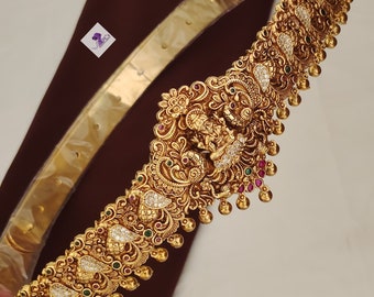 May include: A gold waist belt with intricate floral and paisley designs, featuring a large central pendant with a deity figure. The belt is adorned with clear and red gemstones.