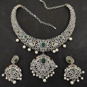 May include: A silver and white necklace set with a peacock design and green stones. The necklace has a chain and a pendant with a peacock design. The earrings are also peacock-shaped with green stones.