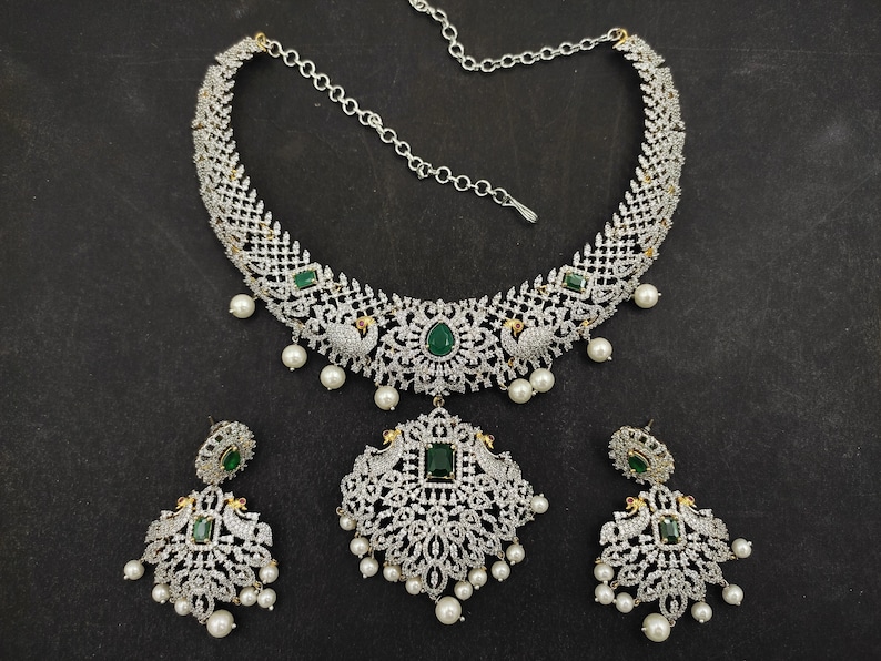 May include: A silver and white necklace set with a peacock design and green stones. The necklace has a chain and a pendant with a peacock design. The earrings are also peacock-shaped with green stones.