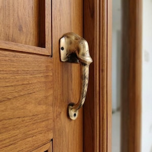 May include: A brass door handle shaped like an elephant head and trunk mounted on a light brown wooden door. The metal handle features engraved details on the trunk and head, with two visible screw holes securing the top plate to the door surface.