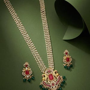 May include: A long necklace featuring five strands of small white pearls and a large gold pendant with a central rectangular red stone, peacock motifs, and green bead accents, accompanied by matching drop earrings with red stones.
