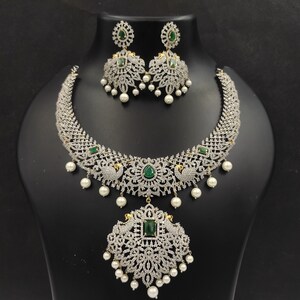 May include: A silver and white necklace set with a large pendant featuring a peacock design and green gemstones. The necklace has a delicate chain and is adorned with small white pearls.