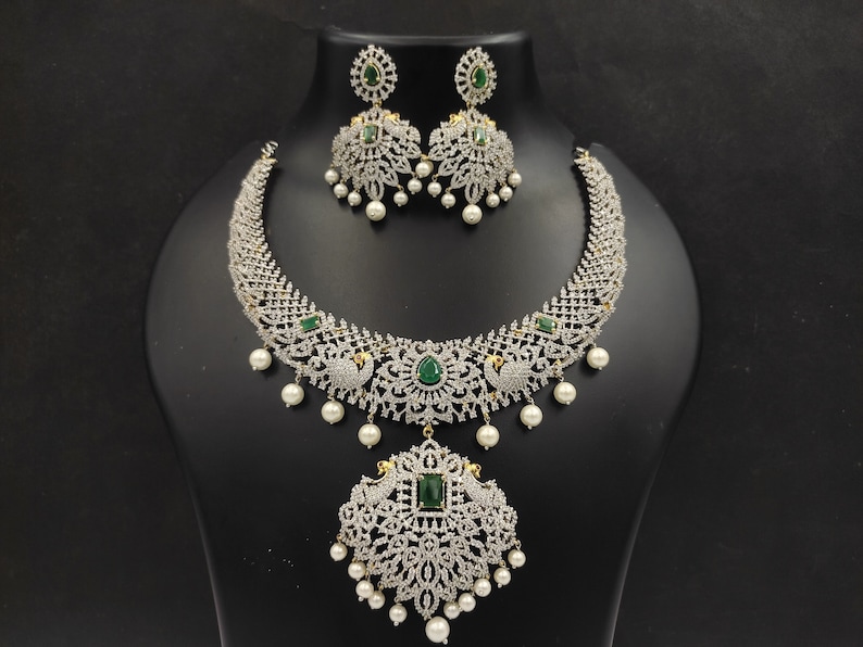 May include: A silver and white necklace set with a large pendant featuring a peacock design and green gemstones. The necklace has a delicate chain and is adorned with small white pearls.