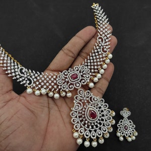May include: A gold necklace with a red stone pendant and white pearl accents. The necklace is made of cubic zirconia and has a delicate design. The necklace is paired with matching earrings.