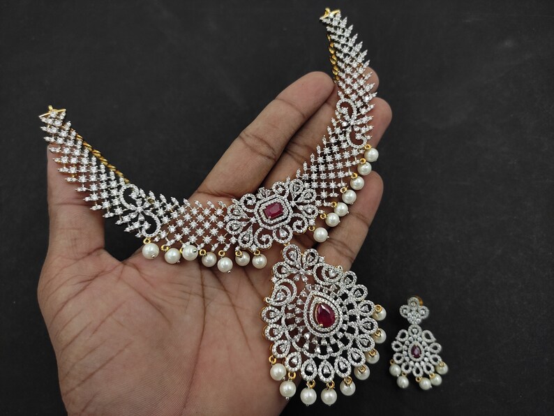 May include: A gold necklace with a red stone pendant and white pearl accents. The necklace is made of cubic zirconia and has a delicate design. The necklace is paired with matching earrings.