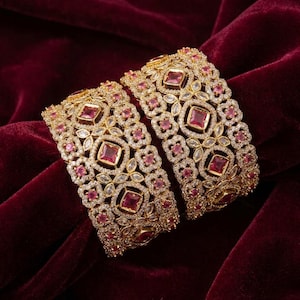 May include: Two wide gold-tone bangles featuring an intricate filigree pattern encrusted with small clear crystals and larger square-cut pink stones. The metalwork displays a repeating geometric design with floral-like motifs throughout the surface.