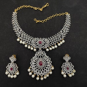 May include: A silver and white necklace set with a large pendant and matching earrings. The necklace features a delicate chain with a large, ornate pendant that is decorated with small, sparkling stones. The earrings are also decorated with small, sparkling stones and have a teardrop shape.