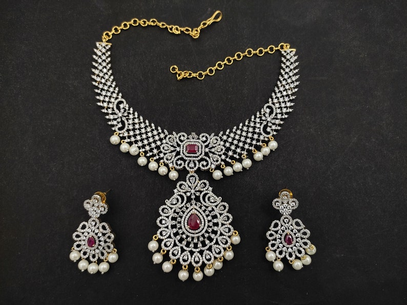 May include: A silver and white necklace set with a large pendant and matching earrings. The necklace features a delicate chain with a large, ornate pendant that is decorated with small, sparkling stones. The earrings are also decorated with small, sparkling stones and have a teardrop shape.
