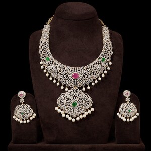 Op de afbeelding: Een sierlijke ketting- en oorbellenset, gepresenteerd op een bruine standaard. De sieraden hebben gedetailleerd zilverkleurig metaalwerk, gezet met heldere stenen en versierd met rode en groene edelstenen. De ketting heeft een hanger met bungelende parels.