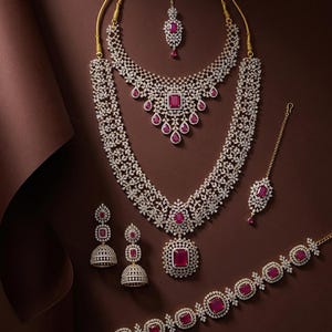 May include: A jewellery set featuring two layered necklaces, a pair of bell-shaped earrings, a bracelet, and two hair accessories. Each piece is crafted with silver-toned metal, clear pavé stones, and rectangular or teardrop-shaped red gems.