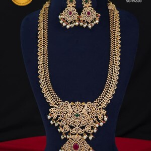 May include: A gold finish bridal haram necklace set with CZ stones, featuring a long chain and a large pendant with peacock motifs, green and red accents, and dangling white pearls. Text reads: PREMIUM QUALITY GOLD FINISH CZ STONES BRIDEL HARAM SGH%590.