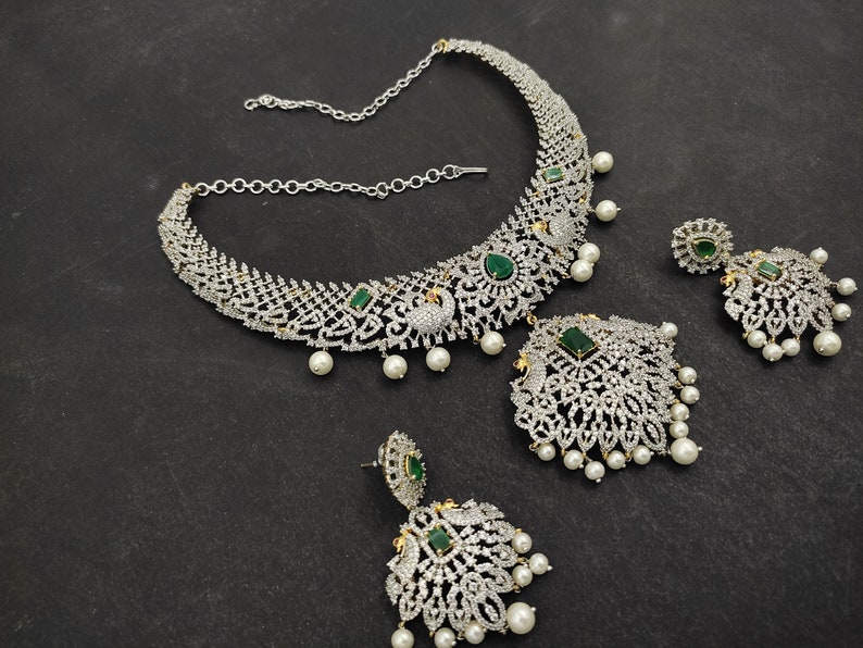 May include: A silver and green necklace and earrings set with a peacock design. The necklace has a chain and a pendant with green stones and white pearls. The earrings are dangle earrings with green stones and white pearls.