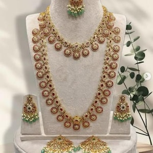 May include: A gold-toned jewelry set featuring a necklace, earrings, and a headpiece. The necklace and earrings have a circular design with red and white accents. The headpiece and earrings have green and white accents. The jewelry is displayed on a mannequin.