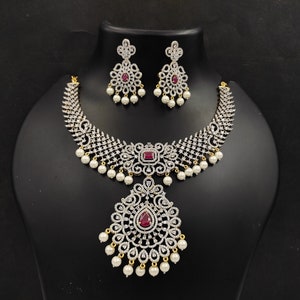 May include: A silver and white necklace with a large pendant and red stones. The necklace is adorned with pearls and has a matching pair of earrings.