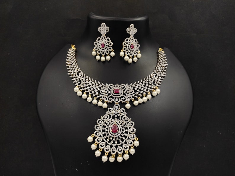 May include: A silver and white necklace with a large pendant and red stones. The necklace is adorned with pearls and has a matching pair of earrings.