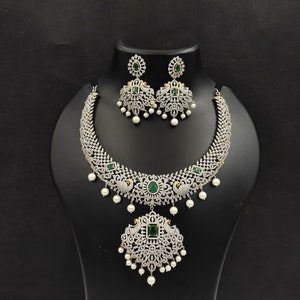May include: A silver necklace and earrings set with a peacock design. The necklace has a large pendant with green stones and white pearls. The earrings have green stones and white pearls.