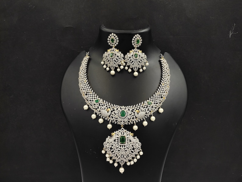 May include: A silver necklace and earrings set with a peacock design. The necklace has a large pendant with green stones and white pearls. The earrings have green stones and white pearls.