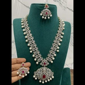 Puede incluir: Un collar plateado y blanco con un gran colgante que presenta una piedra roja y detalles de perlas. El collar se combina con pendientes a juego con una piedra roja y detalles de perlas.