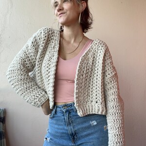 May include: A cream-colored, open-front crochet cardigan. The cropped sweater has long sleeves and a textured stitch pattern. The model is wearing a pink tank top and blue jeans. The cardigan is a versatile piece for layering.