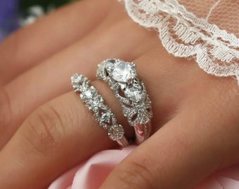 May include: A silver engagement ring set with a large oval-shaped center stone and smaller accent stones. The band is decorated with intricate floral designs.