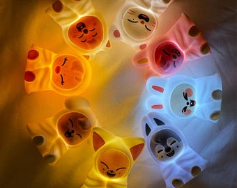 May include: A circular arrangement of seven glowing, cartoon-style night lights. Each light is shaped like an animal, including a cat, bunny, and fox, with a soft, warm glow. The lights are in shades of yellow, pink, and white, creating a whimsical and comforting ambiance.