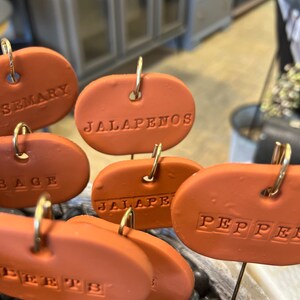 May include: A set of terracotta plant markers with gold wire loops. The markers are oval shaped and have the words "Rosemary", "Jalapenos", "Sage", "Jalapeno", "Pepper", and "Peppers" written on them.