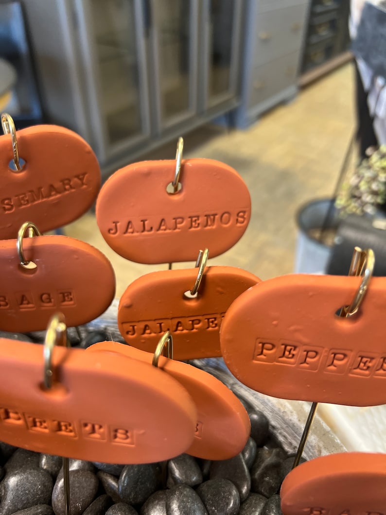 May include: A set of terracotta plant markers with gold wire loops. The markers are oval shaped and have the words "Rosemary", "Jalapenos", "Sage", "Jalapeno", "Pepper", and "Peppers" written on them.