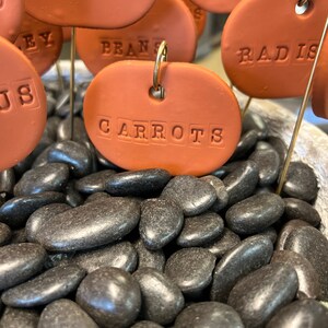 May include: A collection of brown terracotta plant markers with white lettering, including "BEETS", "PEPPERS", "RADISH", "CARROTS", and "BEANS". The markers are placed in a bed of black river rocks.