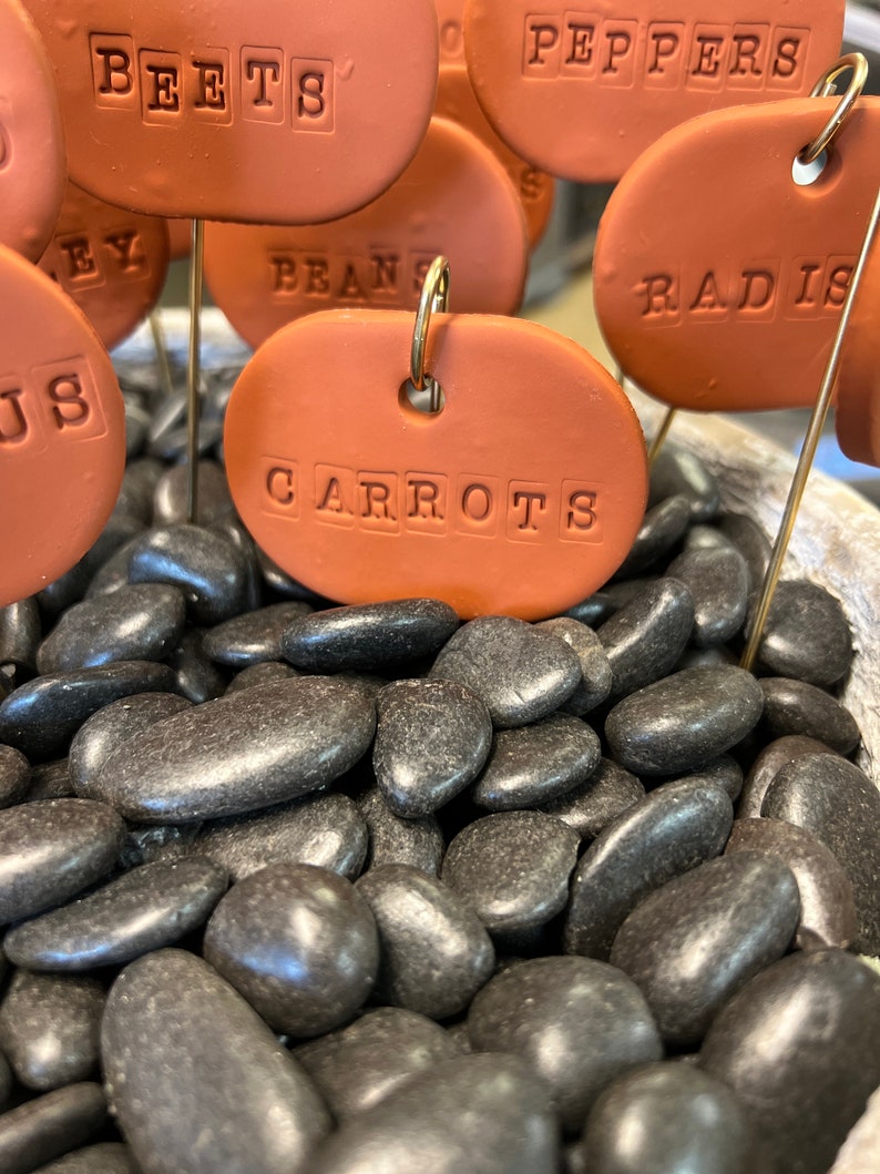 May include: A collection of brown terracotta plant markers with white lettering, including "BEETS", "PEPPERS", "RADISH", "CARROTS", and "BEANS". The markers are placed in a bed of black river rocks.