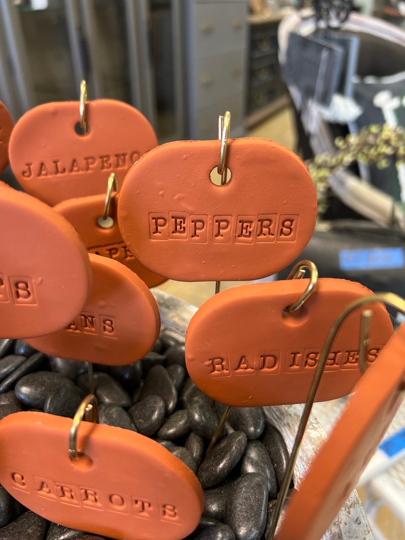 May include: Terracotta plant markers with the words "Jalapeno", "Peppers", "Radishes", and "Carrots" written on them. The markers are on a bed of black pebbles.