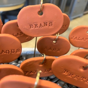 May include: A set of terracotta plant markers with metal rings and wire stems. The markers are oval shaped and have the words "BEANS", "SAGE", "PARSLEY", "BEETS", "JALAPENO", "CILANTRO", and "AGUS" written on them.