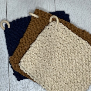 May include: Three crocheted pot holders in navy blue, brown, and beige with wooden rings for hanging.