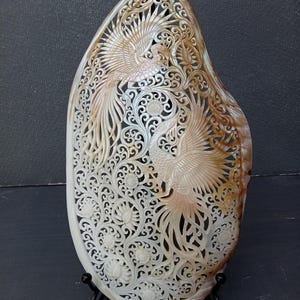Könnte beinhalten: Eine oval geformte Muschelschnitzerei mit komplizierten Durchbrüchen von zwei Phönixvögeln, umgeben von detaillierten Blumen- und Rankenmustern. Die natürliche Muschel zeigt einen Verlauf von cremeweißen bis hin zu warmen Brauntönen auf der Oberfläche.